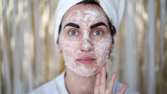 young woman with towel on her head, with cosmetic mask on her face and with cosmetic paint on her eyebrows. Woman puts herself in order, facial skin care and eyebrow correction