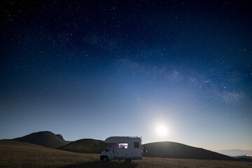 Fototapeta na wymiar Panoramic night sky over Campo Imperatore highlands, Abruzzo, Italy. The Milky Way galaxy arc and stars over illuminated camper van. Camping freedom in unique hills landscape.