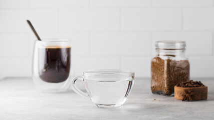 On the grey table there is a mug of water, a mug of black coffee and a jar of instant coffee.