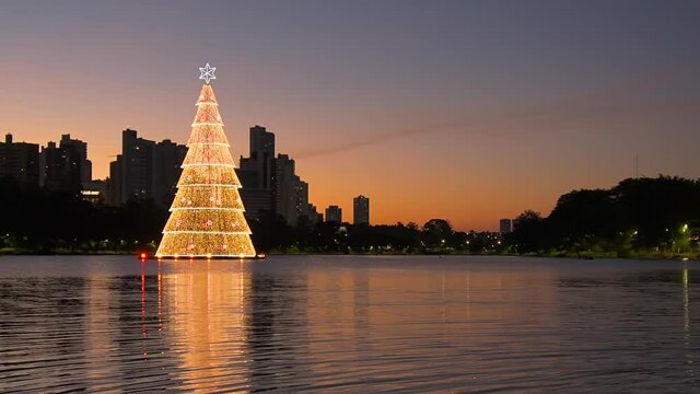 Beautiful city lake at dusk with christmas tree floating, some buildings around and the water of the lake reflecting the buildings.Video at Igapo Lake, Londrina city, Parana state, Brazil. Zoom out.