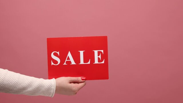 Profile side view closeup woman hand holding red sale card, mega shopping, black friday, huge discounts in mall. Indoor studio shot isolated on pink background.