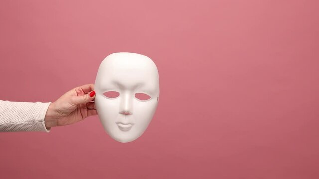Profile side view closeup of woman hand holding white facial mask model, hiding real personality, anonymity. Indoor studio shot isolated on pink background.
