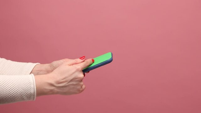 Profile side view closeup of woman hand holding mobile phone in hand, crolling online, surfing internet website. Indoor studio shot isolated on pink background.