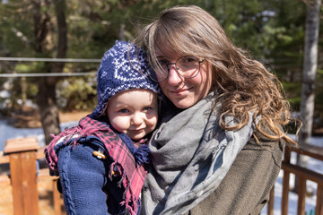 Obraz premium Closeup portrait of thirty year old mom carrying cute three year old toddler wearing blue wooly hat outdoors in cold weather with blurred background.