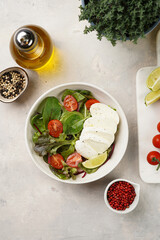 Salad with green leaves, lettuce, spinach, cherry tomatoes, mozzarella, olive oil, chilli pepper, lime juice on grey stone surface, top view