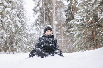 Fototapeta na wymiar Happy teenager boy sitting on snow in winter forest. Child having fun outdoors. Joyful adolescent playing in snow at snowfall. Laughing smiling kid walking in winter park in cold weather