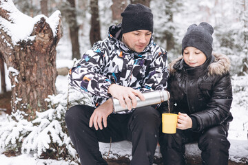 Father and sons drinking tea from thermos and talking sitting together on log in winter snowy...