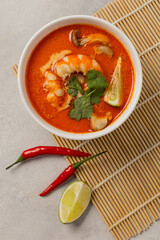 White bowl of fresh Tom Yum soup on a bamboo stand with ingredients on a light background