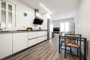 Long kitchen with white furniture and gray countertop, dining table and tv in the background