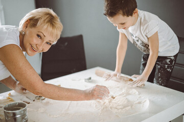 A grandmother and grandson are cooking cookies in the kitchen, they smile and roll out the dough on...