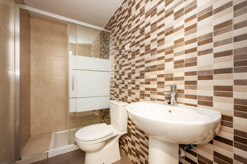 Toilet with white porcelain sanitaryware, glass partition with aluminum frames and tiled wall in brown and cream colors