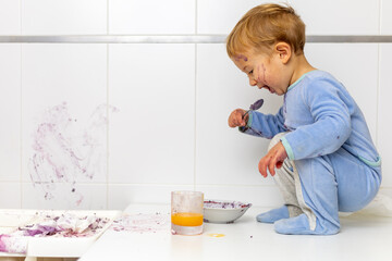 niño rubio en pijama azul en cuclillas sobre la mesa tomando su desayuno de arandanos con la pared...