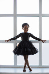 African american little kid wearing stylish black dress standing in some ballet pose near window at...