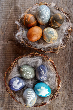 Vertical image of eggs colored with natural dyes and accented with botanical leaf and flower prints, nestled into fluffy gray alpaca fiber in a woven grapevine nest displayed on a weathered gray board