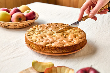 Hand of woman cutting slice of homemade apple cake Charlotte or american sponge apple pie with walnuts and ripe red apples on the table with linen tablecloth. Healthy seasonal eating.