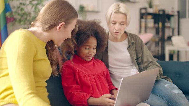 Little black girl browsing internet on laptop together with foster lgbt parents
