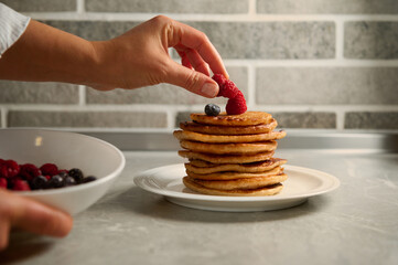 The chef's hand puts raspberries on a stack of delicious homemade freshly baked pancakes served on...