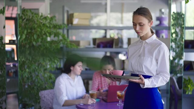 Portrait of tired young waitress standing on the right in cafe with blurred happy clients at background. Exhausted overworking Caucasian woman posing indoors in slow motion. Employment concept