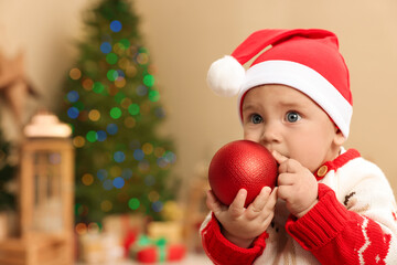 Fototapeta na wymiar Cute little baby in sweater and Santa hat with Christmas bauble at home, space for text. Winter holiday