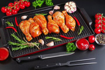Raw chicken legs marinated in red sauce in a grill pan on a dark background with herbs, red hot pepper tomatoes and garlic. Chicken half-cooked legs. Top of view.