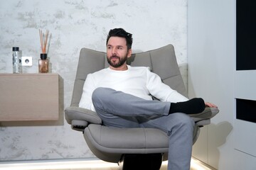 Portrait of handsome successful rich relaxed man, young person is sitting in comfortable armchair at his luxury flat, apartment or home, relaxing, resting, having a rest, dreaming, thinking.