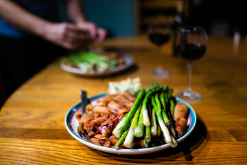 Closeup of steamed asparagus stems on two plates with red wine tomato sauce rigatoni pasta in marinara with dimmed light romantic atmosphere and woman plating setting table in background