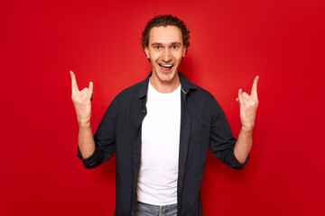 excited man with his mouth open shows rock sign with his hands depicting listening to his favorite music heavy metal. isolated on red background. concept - people, symbols, rock and roll, freedom