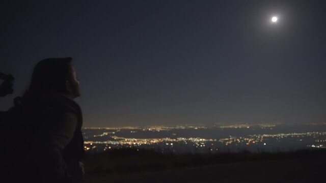 Woman with a tripod and backpack walking under the moonlight, city light in the background. 