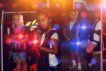 Fototapeta na wymiar Portrait of glad african tweenager girl with laser pistol posing in laser tag labyrinth on background with her team
