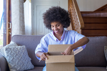 African woman sitting on sofa in living room hold on lap big carton box unpacking parcel received...