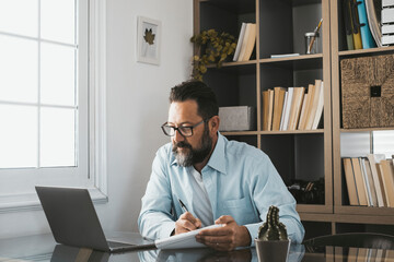 Bearded handsome adult male looking at laptop screen reading email or content and writing notes at home office. Focused businessman using laptop while writing down notes in modern office