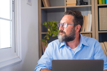 Serious bearded caucasian man in deep thoughts looking out through office window. Businessman taking a break from work and admiring view from workplace. Male executive looking away while working 