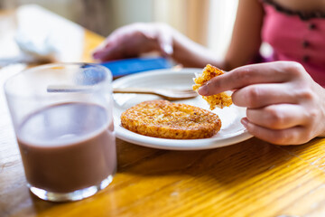Macro closeup of woman sitting at breakfast table holding eating potato fried hash browns with hand on plate by glass of chocolate milk and background of phone