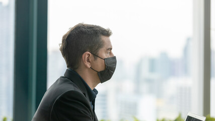 A businessman in black dress wearing face mask is using laptop to communicate with friends and...