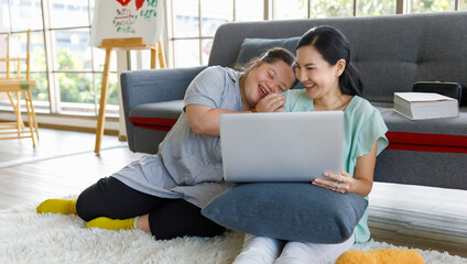 Portrait shot of Asian lovely mother sitting on floor and young chubby down syndrome autistic autism little daughter lay down on cozy sofa smiling look at camera watching cartoon from laptop computer