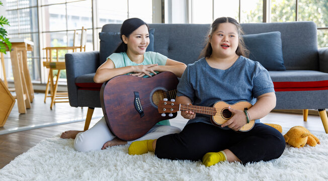 Asian Lovely Mother Holding Guitar Sit Smiling On Sofa Couch In Living Room At Home Teaching Young Chubby Down Syndrome Autistic Autism Little Daughter Playing Ukulele And Singing Music Song Lesson