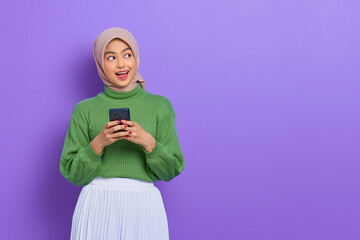 Shocked beautiful Asian woman in green sweater and hijab using a mobile phone and looking aside isolated over purple background