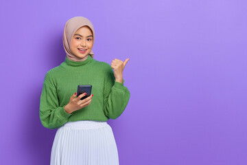 Portrait of smiling beautiful Asian woman in green sweater holding mobile phone and pointing finger aside isolated over purple background