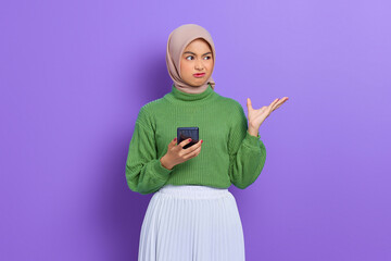 Confused beautiful Asian woman in green sweater and hijab using mobile phone isolated over purple background