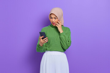 Amazed beautiful Asian woman in green sweater and hijab using a mobile phone, reading good news message isolated over purple background