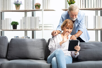 senior couple, elderly man and woman using tablet and say hello to someone on sofa