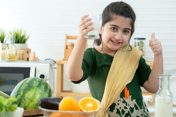 Smiling little Indian girl holding a glass of milk and thumb up in kitchen at home, healthy concept