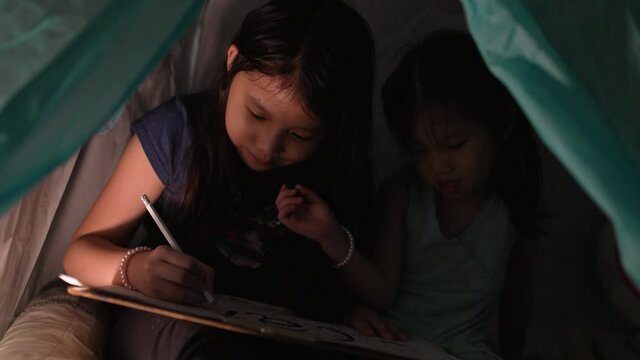 Asian girl with her sister drawing picture and play in teepee tent with flash light at night in bedroom. Staycation concept