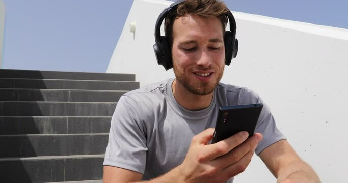Podcast. Man listening to podcasts or audiobook on mobile phone app wearing headphones sitting outdoors on stairs. Healthy lifestyle sport athlete using smartphone on break outsite relaxing