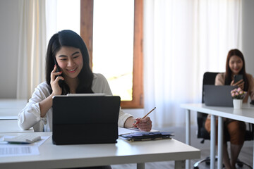 Cheerful asian female office worker talking on mobile phone and working with computer tablet.