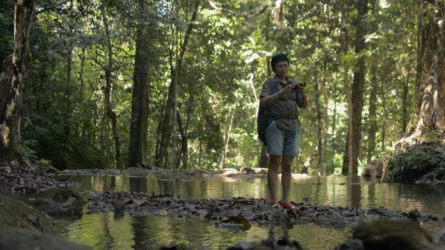 Asian female hiker with backpack takes a photo with mobile phone in tropical forest while standing among small creek during summer. Travel explorer. Jungle adventure. Solo camping in Thailand.