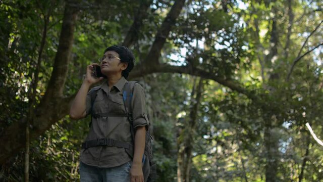 Asian female trekker with backpack using mobile phone talking to family in the tropical forest during summer. Signal network for comprehensive communication. Wireless connectivity technology.