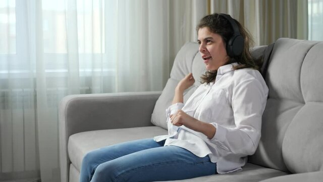 Disabled woman in headphones having infantile cerebral paralysis listens to music dancing and singing sitting on sofa in living room at home.
