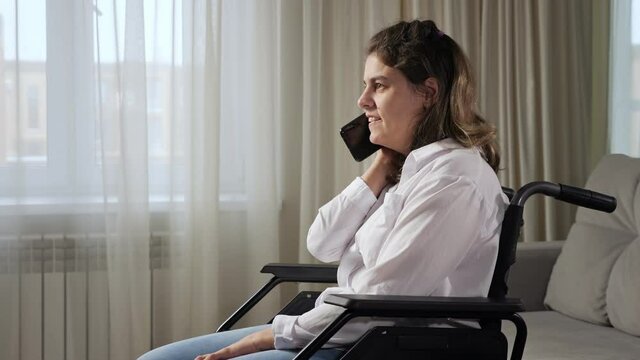 Disabled woman with infantile cerebral paralysis talks smiling on mobile phone sitting in wheelchair near window in living room at home side view.