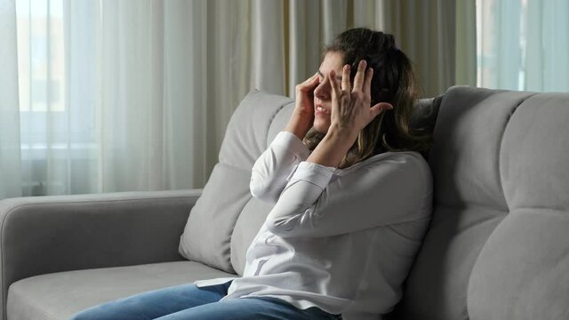 Disabled woman with organic damage of central nervous system suffers from strong headache sitting on grey sofa near window at home side view.